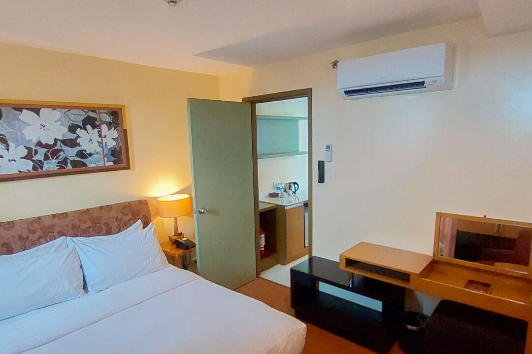 A hotel room with a bed, bedside table, lamp, air conditioning unit, and a small desk with a mirror.