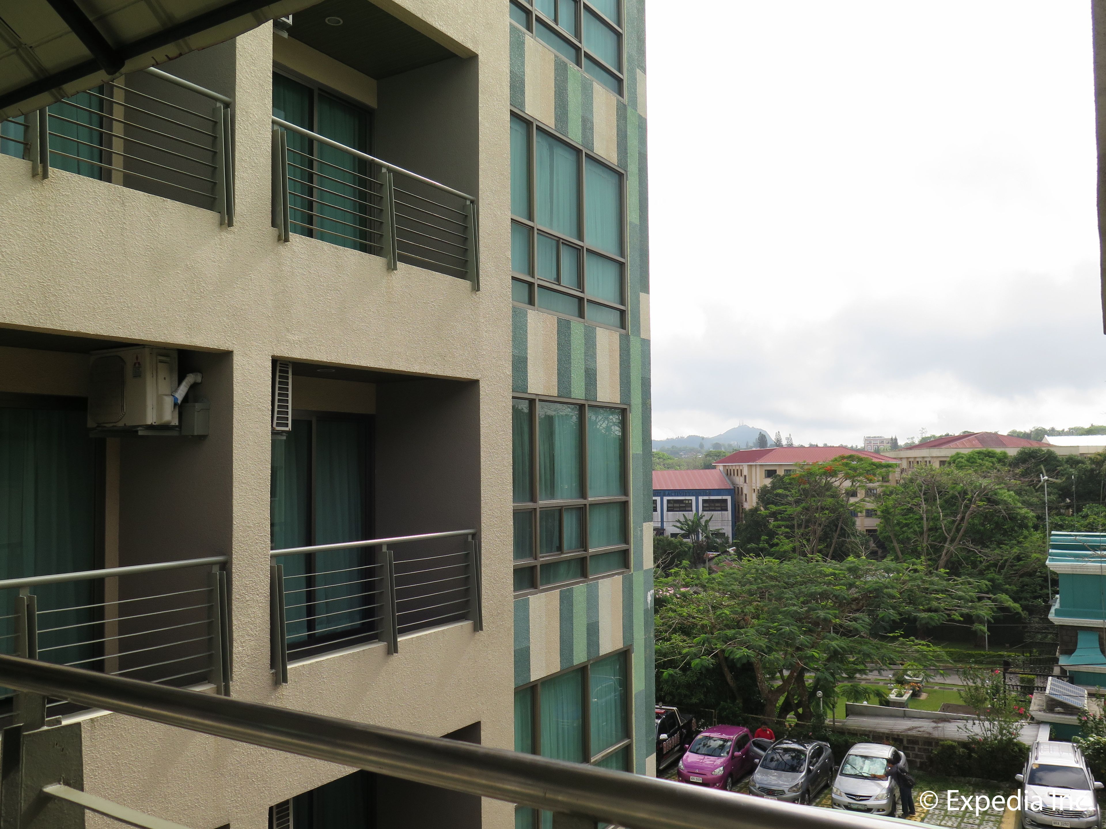 Un edificio de varios pisos con balcones, vista a vehículos estacionados y vegetación circundante.