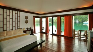 A bedroom with a large bed, wooden flooring, a view of a lake, and a balcony with orange curtains.