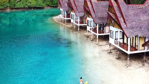 Overwater bungalows with thatched roofs on a clear turquoise beach, surrounded by lush greenery.