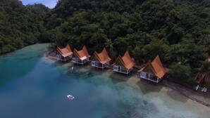 An aerial view of a resort with bungalow-style accommodations with red roofs, situated on a clear turquoise beach surrounded by dense forest.