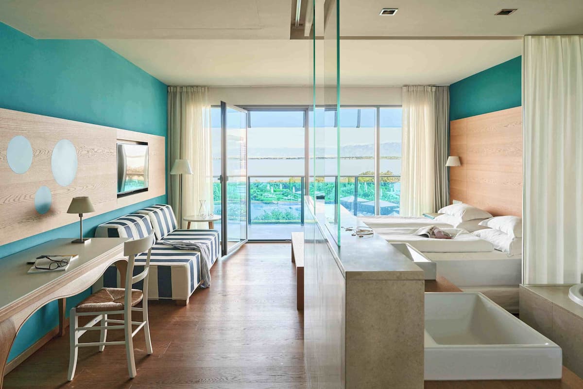 Een moderne hotelkamer met een ruim balkon, een bureau met stoel, een sofa en een afzonderlijk slaapgedeelte met een bad.