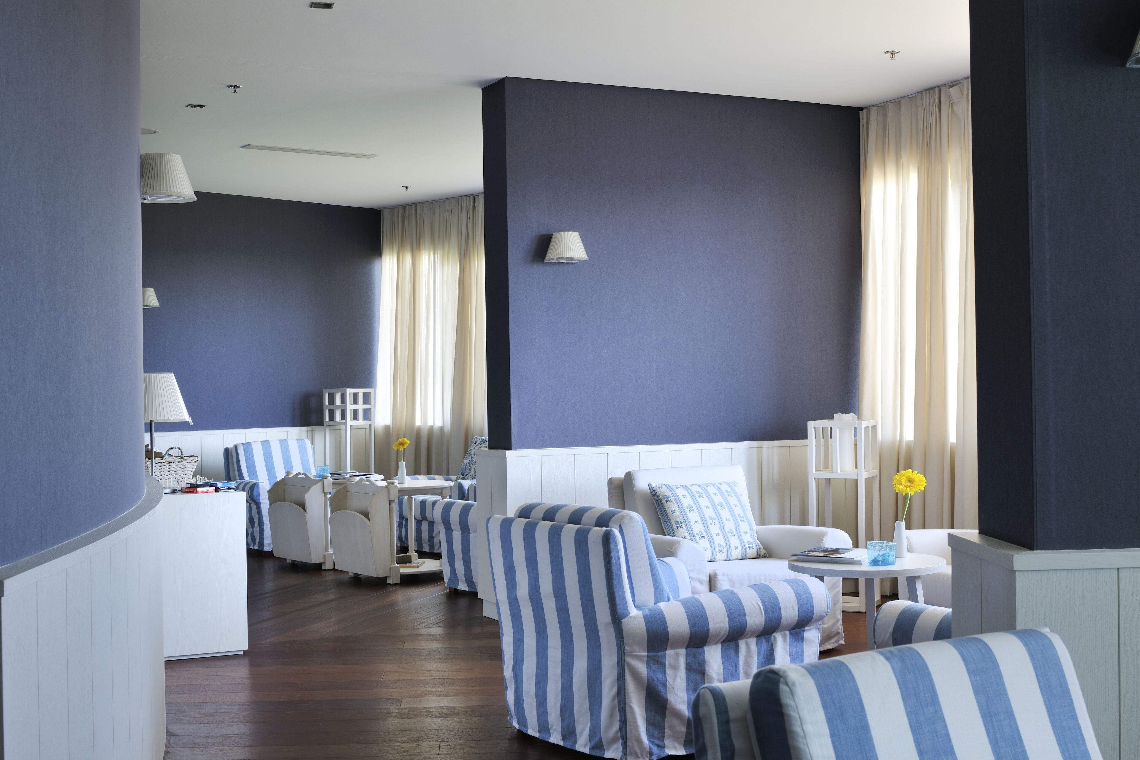 Ein Hotelzimmer mit dunkelblauer Akzentwand, einem weißen Bett, gestreiften Sessel und einem kleinen runden Tisch. Ein Fenster mit Vorhängen ist ebenfalls vorhanden.