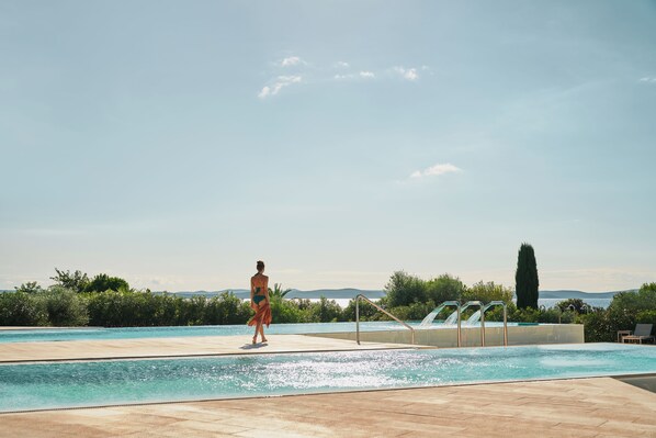 A pool area with a clear view of the sea, a person in a swimsuit, and a poolside lounge area.