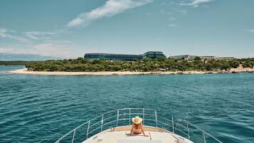 Un bote con una persona en la cubierta, una playa de arena y edificios al fondo.