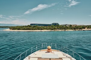 Un bote con una persona en la cubierta, una playa de arena y edificios al fondo.