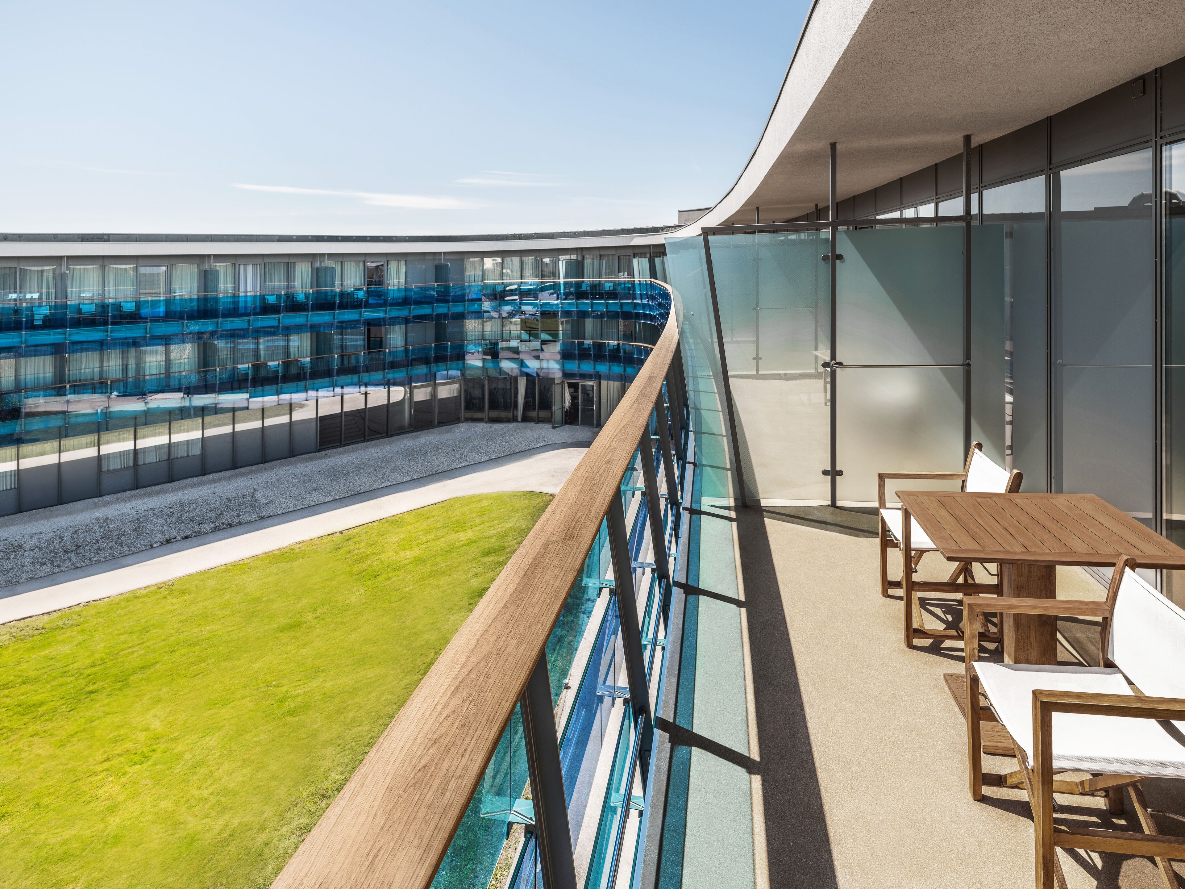 A modern hotel balcony with a glass railing, a pool, and outdoor seating.
