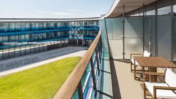 A modern hotel balcony with a glass railing, a pool, and outdoor seating.