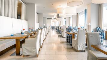 A modern hotel dining area with long tables, white cushioned seating, and large pendant lights.