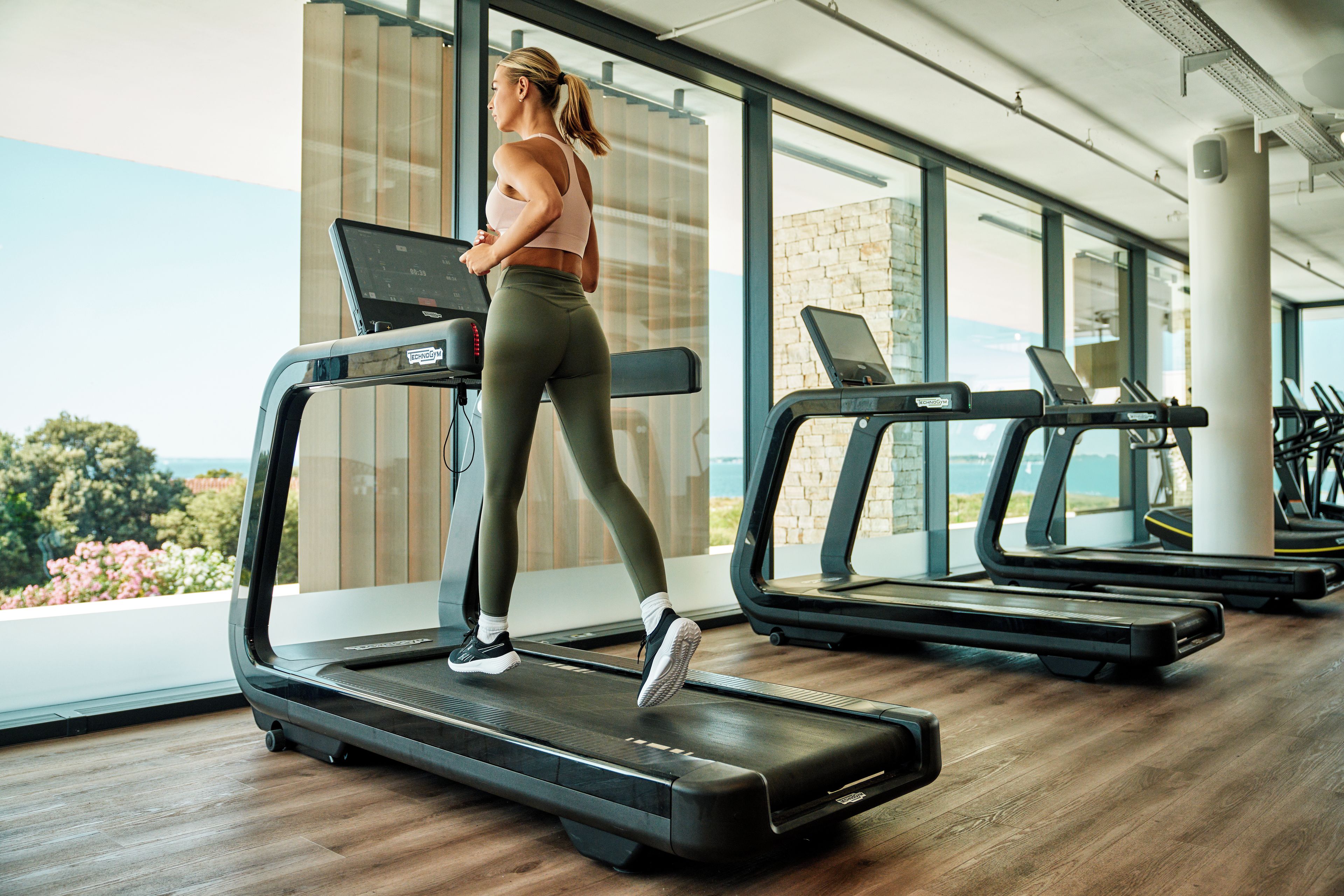 Una mujer usando una cinta de correr en un gimnasio con varias cintas y vistas al exterior.