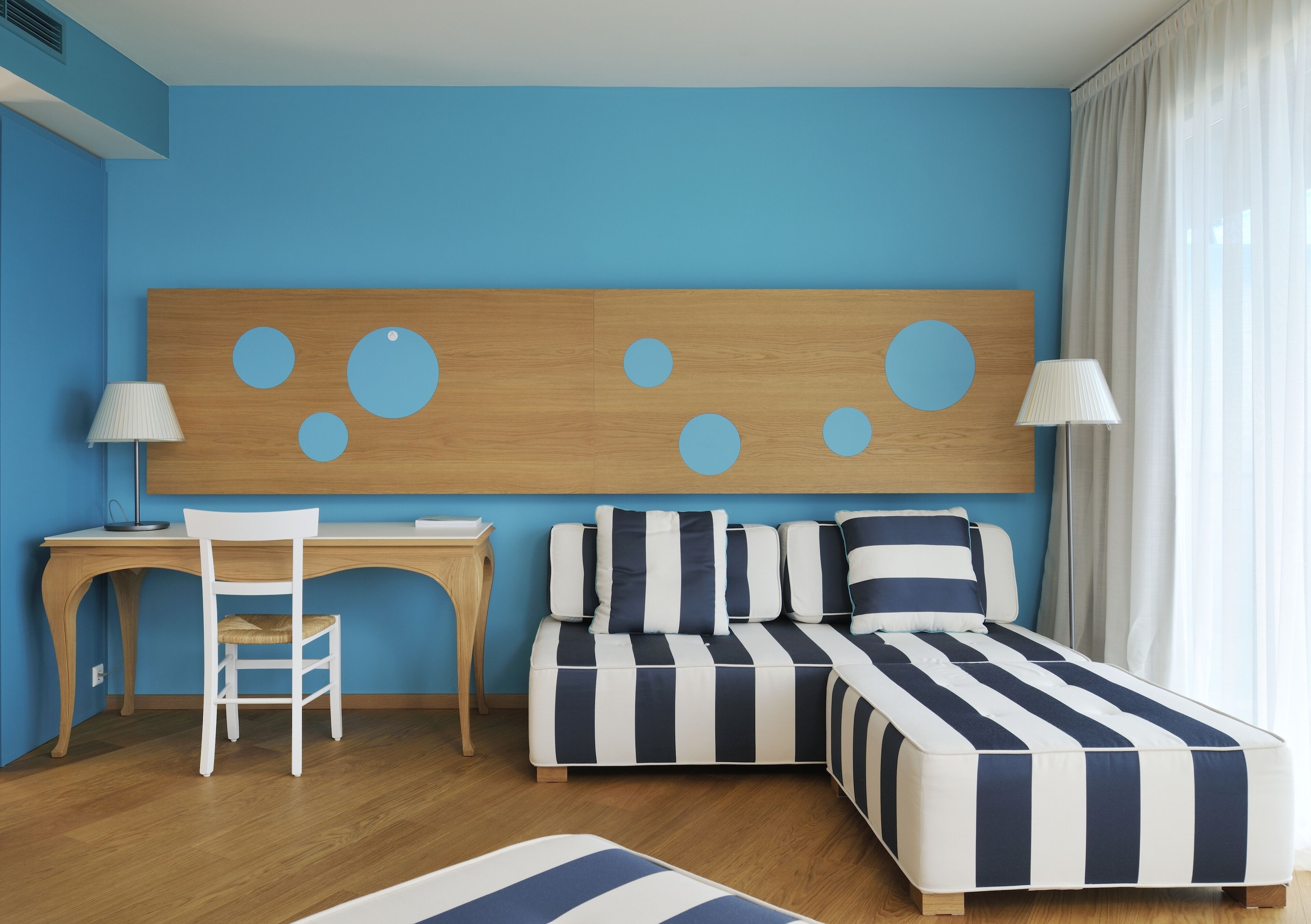 A room with a desk, a chair, a bed with striped bedding, a sofa, and a large wall decoration.