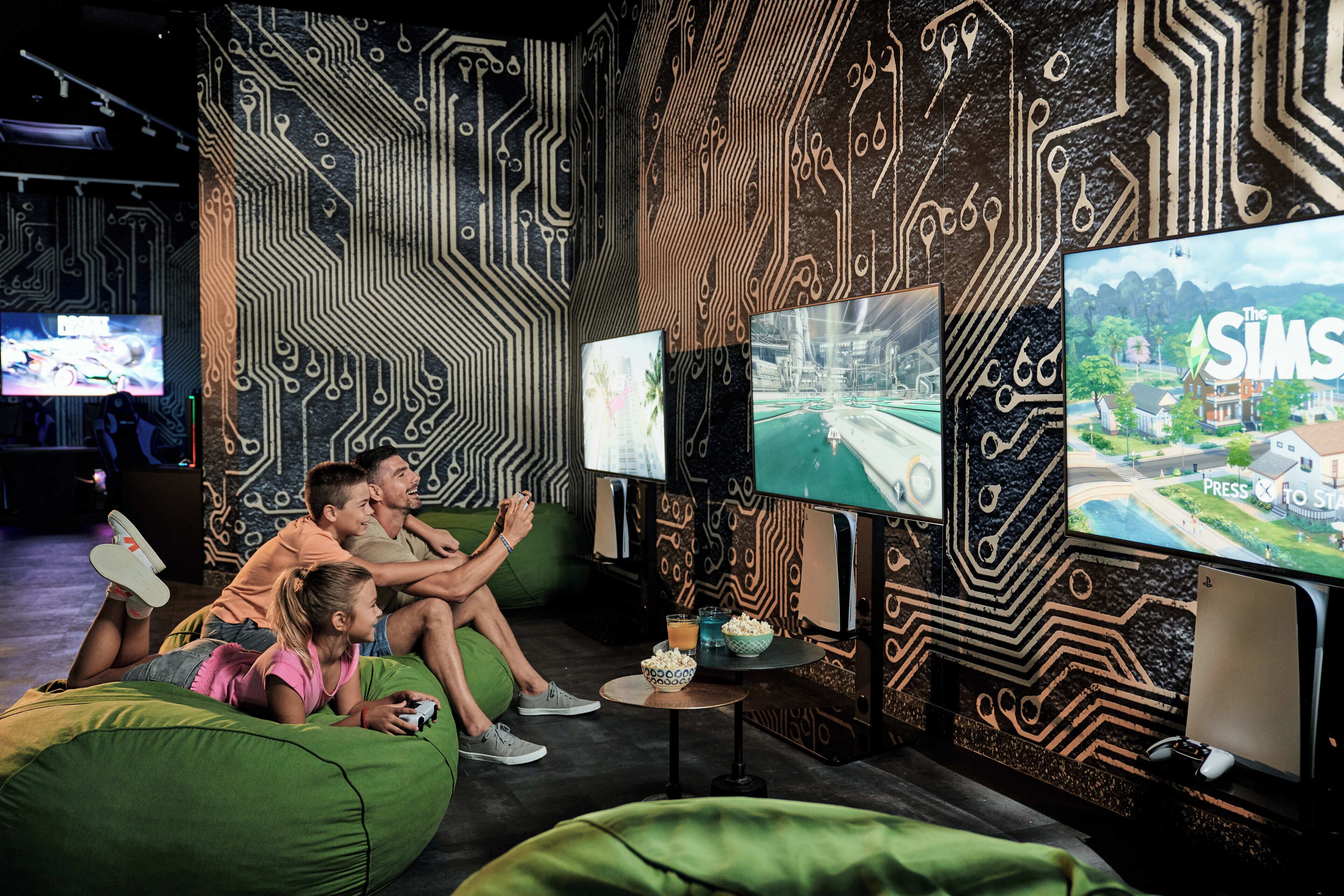 Ein Zimmer mit Wandgestaltung im Stil einer Platine, mehreren Bildschirmen, die Spiele zeigen, und einer Sitzecke mit grünen Sitzsäcken.