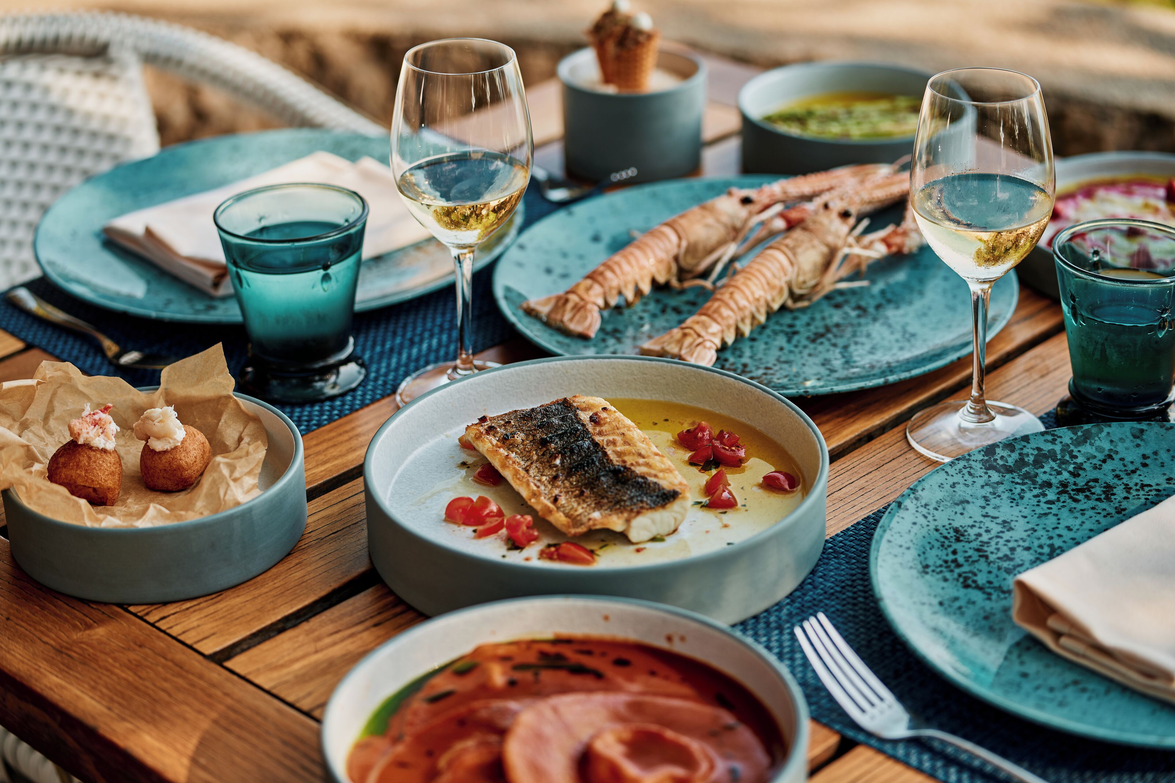 Una mesa de comedor con varios platos, incluyendo mariscos y un plato de pescado, acompañada de copas de vino blanco.