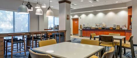 A hotel dining area with multiple tables and chairs, a counter with food and beverage options, and large windows providing natural light.