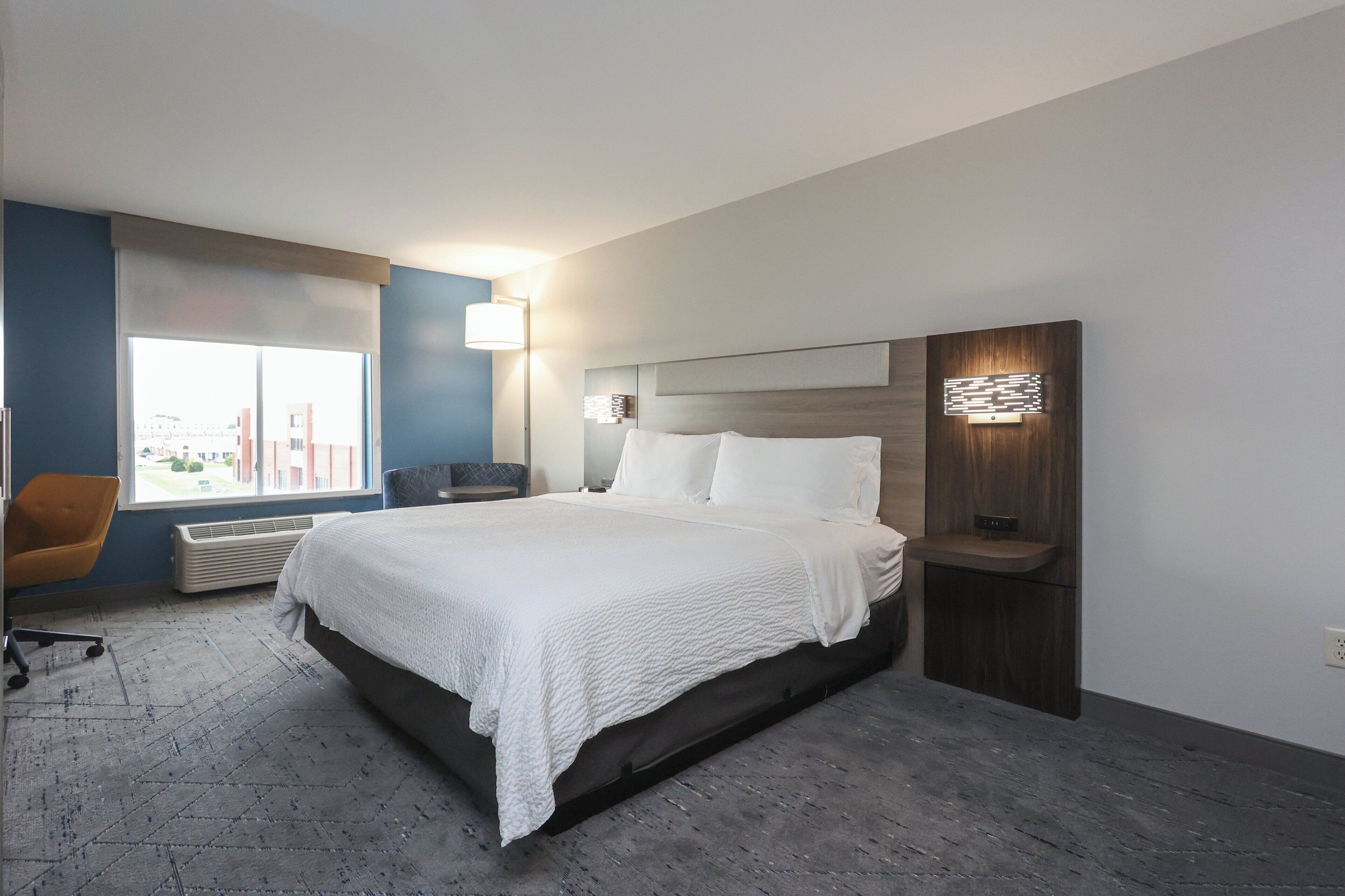 A modern hotel room with a large bed, a desk, a chair, and a view of buildings outside the window.