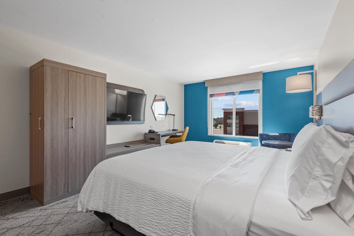 A hotel room with a large bed, a desk, a chair, a wardrobe, a mirror, and a window with a view.