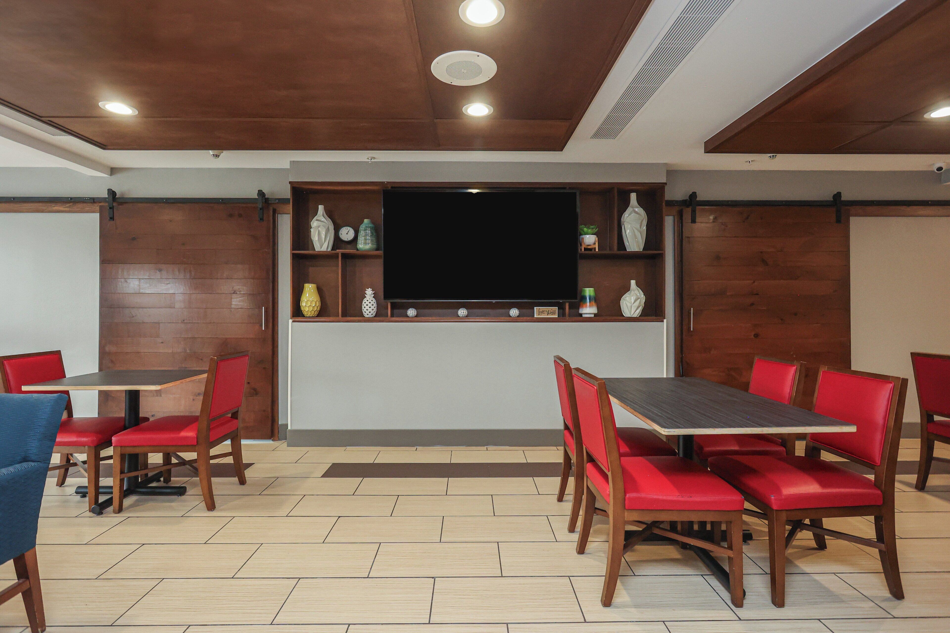 A dining area with wooden tables and red chairs, a flat-screen TV, and decorative items on shelves.