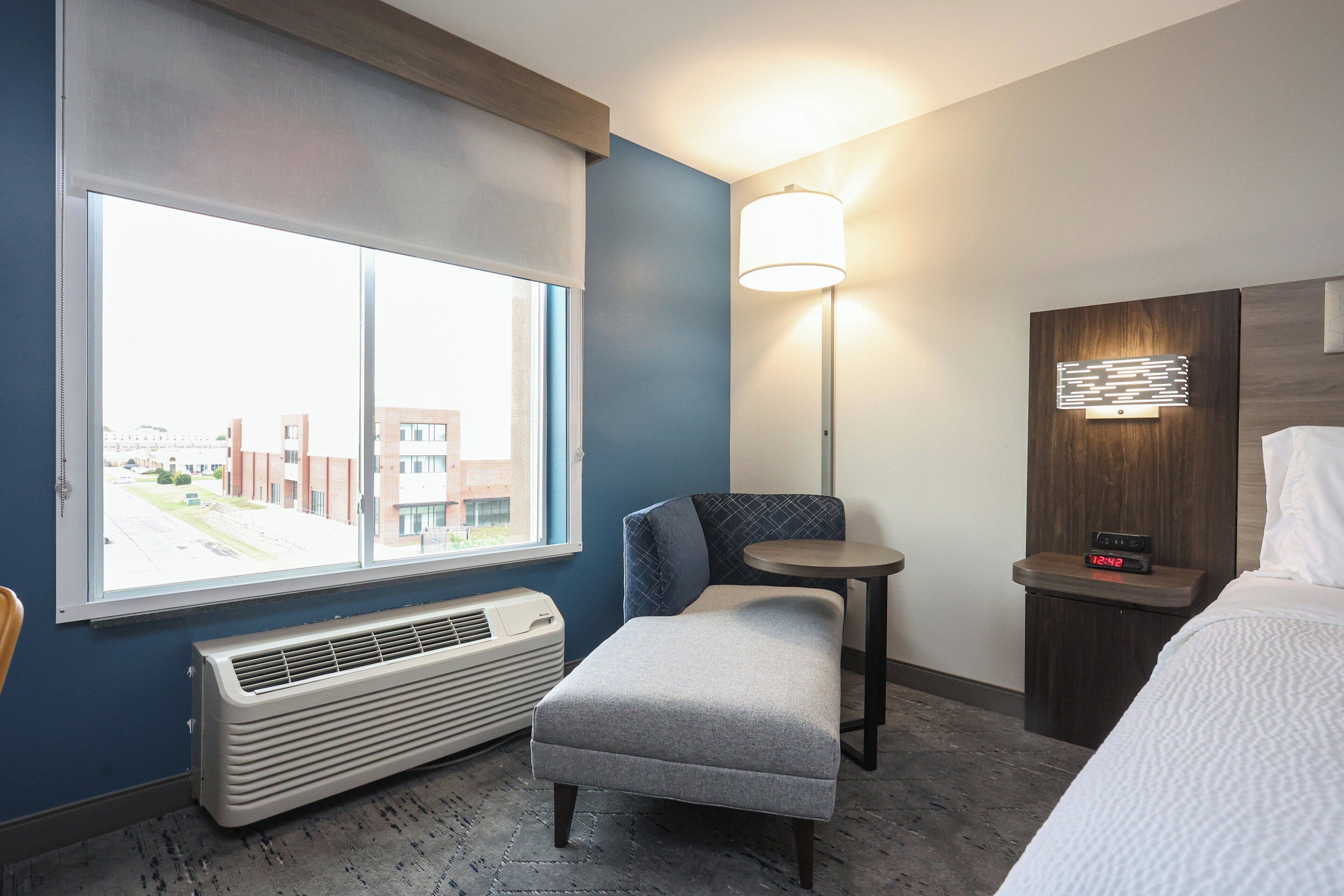 A hotel room with a large window, a standing heater, a small bench, a bedside table with a lamp, and a bed.