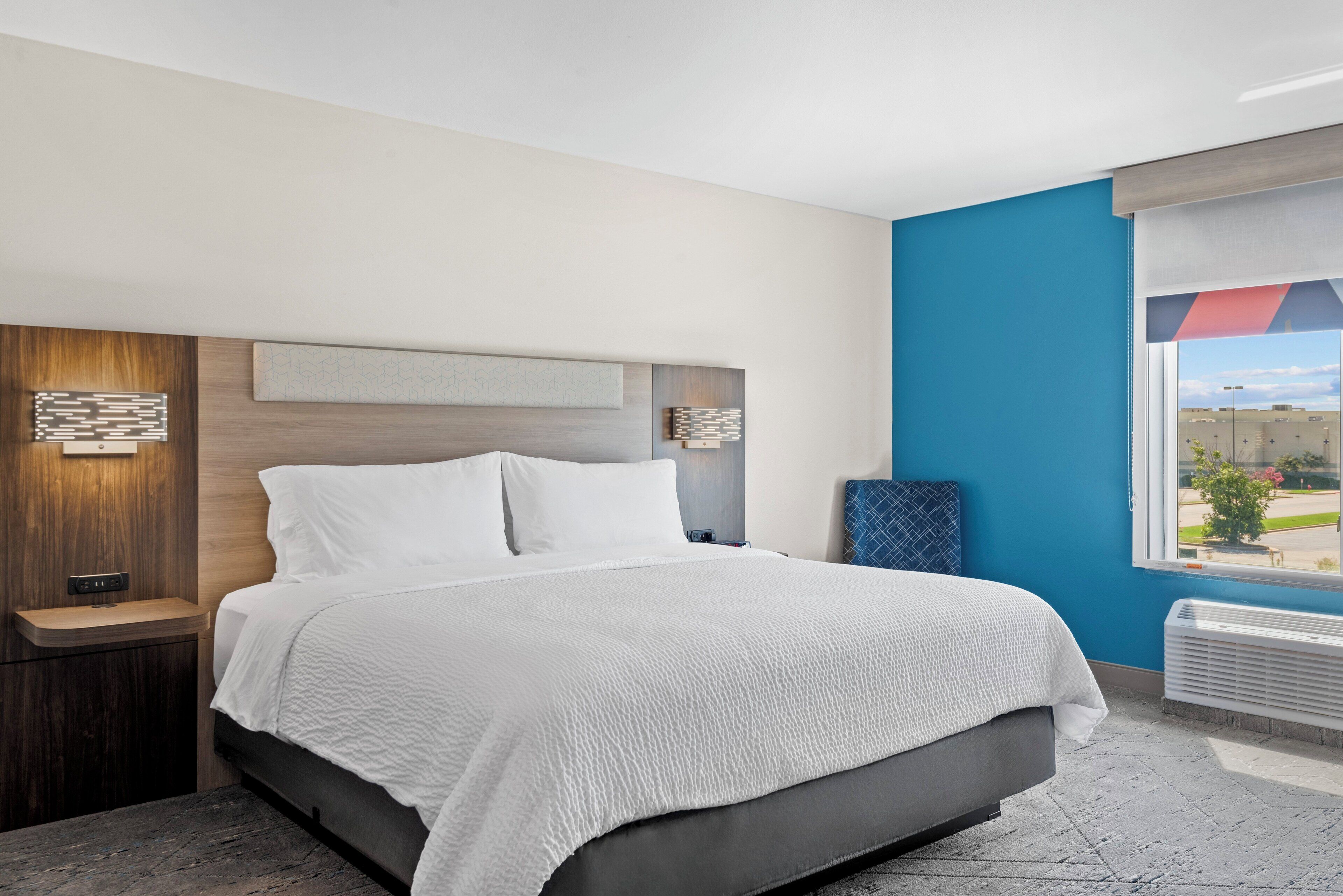 A modern hotel room with a large bed, a nightstand, a wall-mounted light, and a window with a view of the outside.