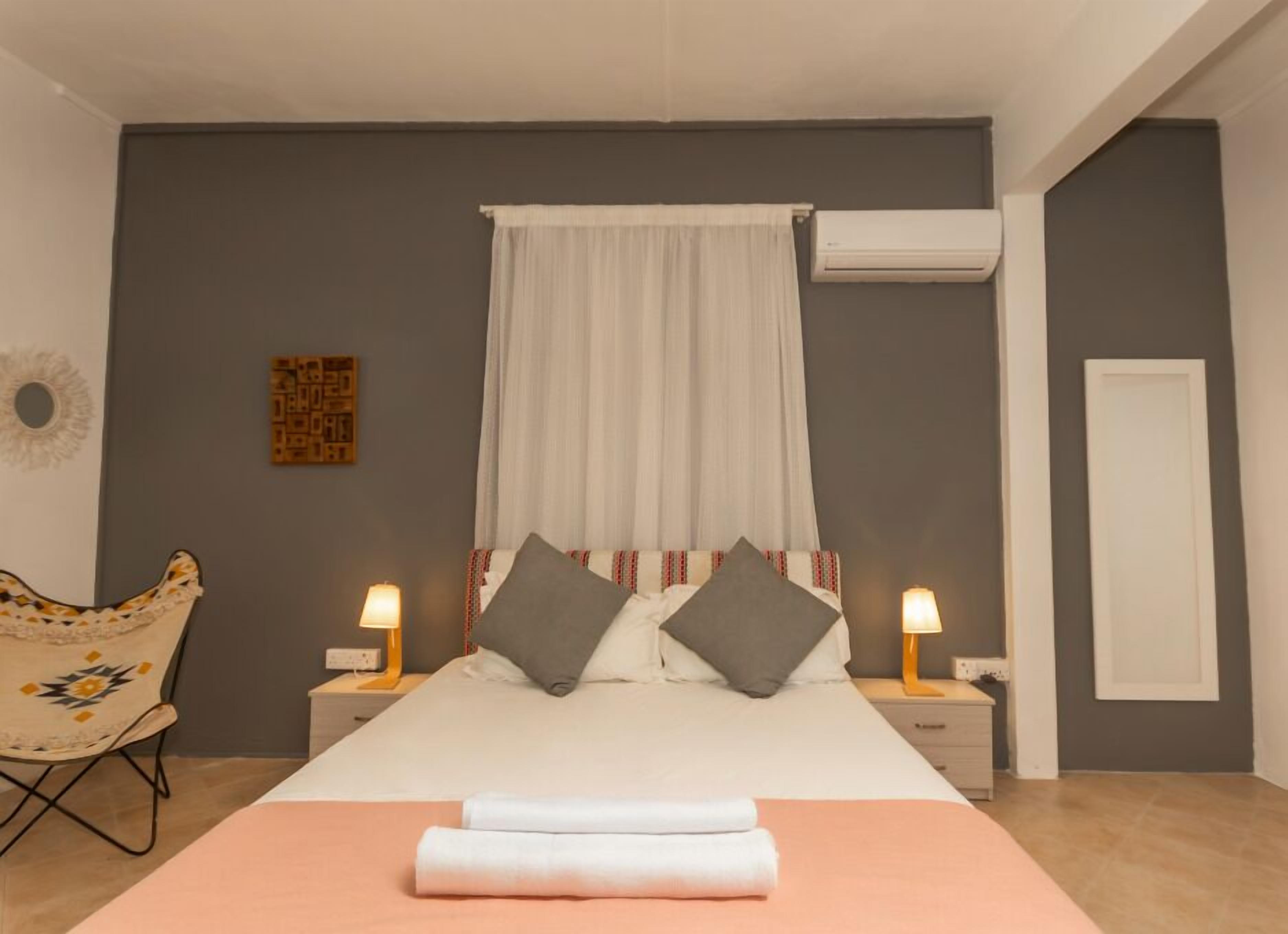 A bedroom with a bed, two bedside lamps, a chair, an air conditioning unit, and a door.