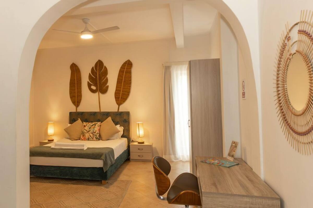 A bedroom with a bed, bedside lamps, a wooden headboard, a mirror, and a wall with decorative leaves.