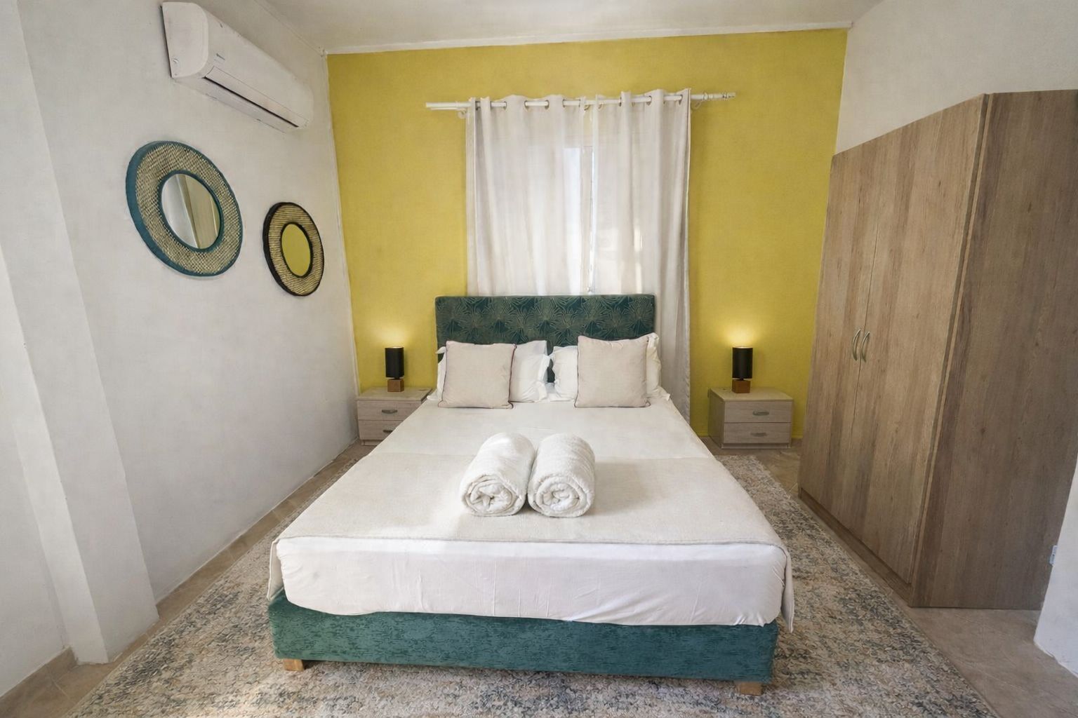 A neatly made bed with two rolled towels, a green upholstered headboard, white bedding, and a yellow accent wall.