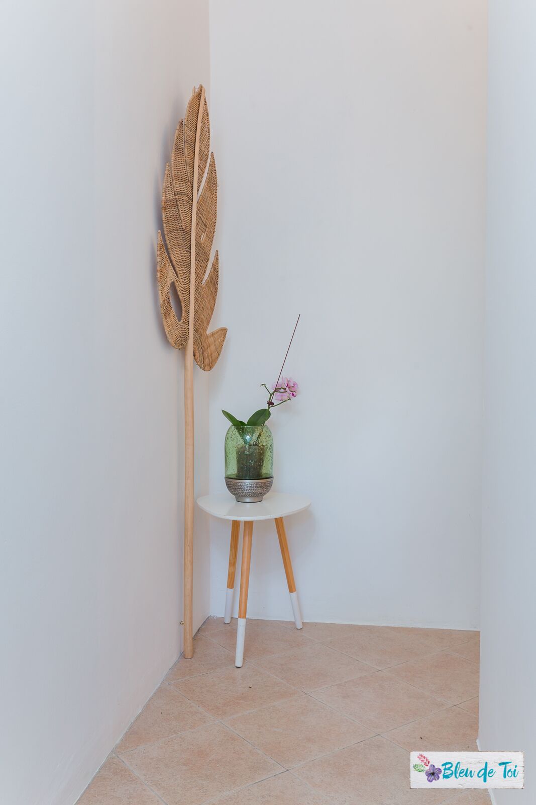 Eine Ecke mit einer weißen Wand, einem kleinen runden Tisch mit Topfpflanze und einer großen gewebten Blattdeko.