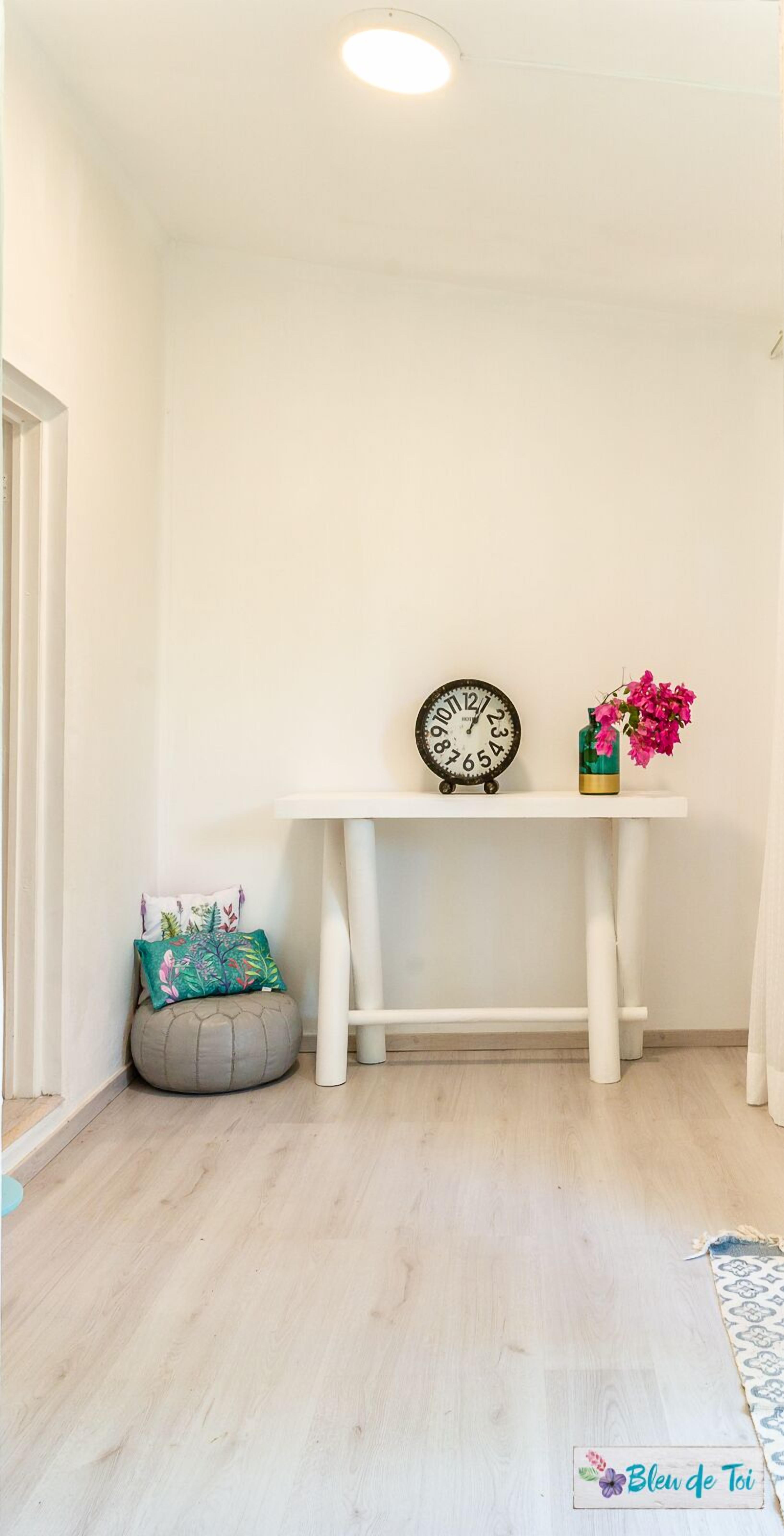 Rincón de una habitación con una consola blanca, un reloj, un jarrón con flores, un sillón informal y una alfombra estampada.