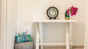 Rincón de una habitación con una consola blanca, un reloj, un jarrón con flores, un sillón informal y una alfombra estampada.