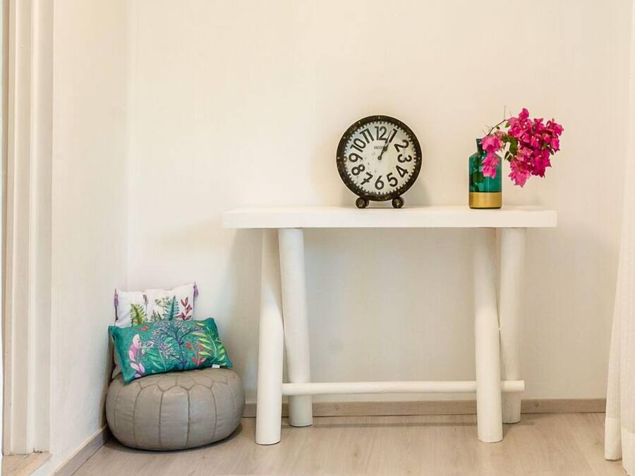Rincón de una habitación con una consola blanca, un reloj, un jarrón con flores, un sillón informal y una alfombra estampada.