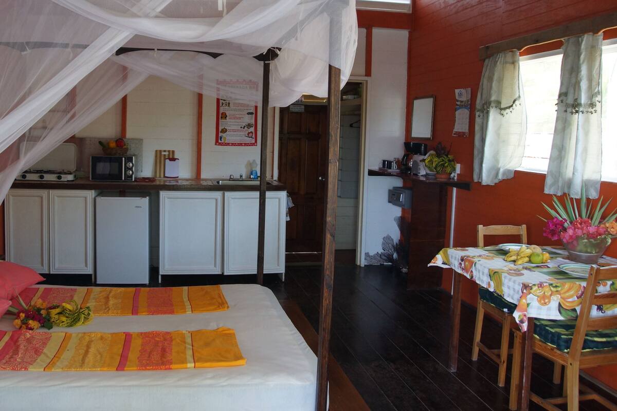 Quarto com cama, mesa de jantar e cadeiras, área de cozinha com armários e mosquiteiro sobre a cama.