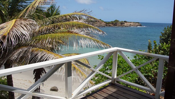 A wooden deck with a white railing overlooking a beach with clear blue water and a rocky outcrop in the distance.
