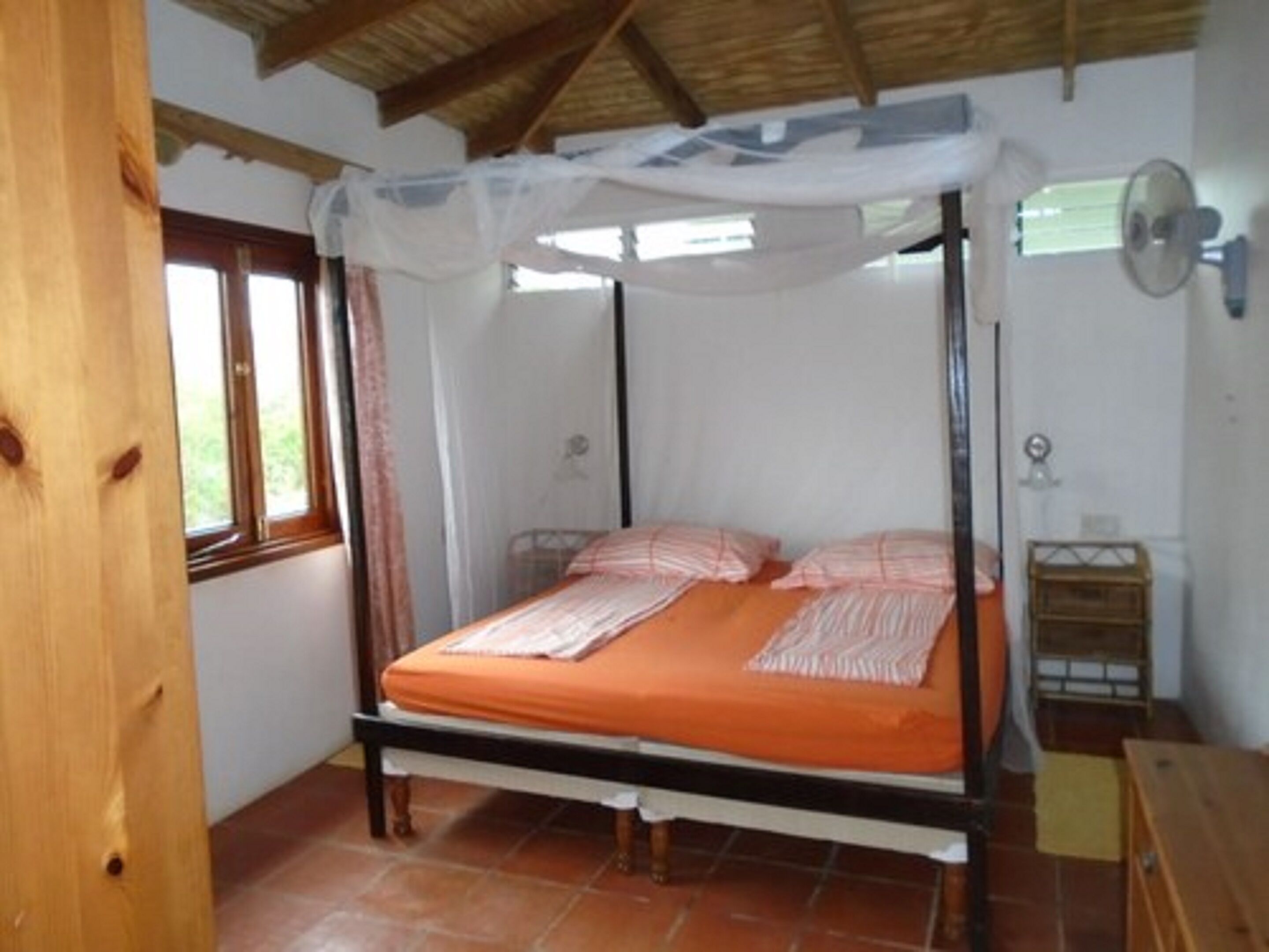 Una cama con dosel sobre cuatro soportes, con una colcha naranja, cabecera de madera y dosel blanco.