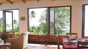 A dining area with a table set for two, large windows with a view of palm trees and greenery, and a cushioned chair.