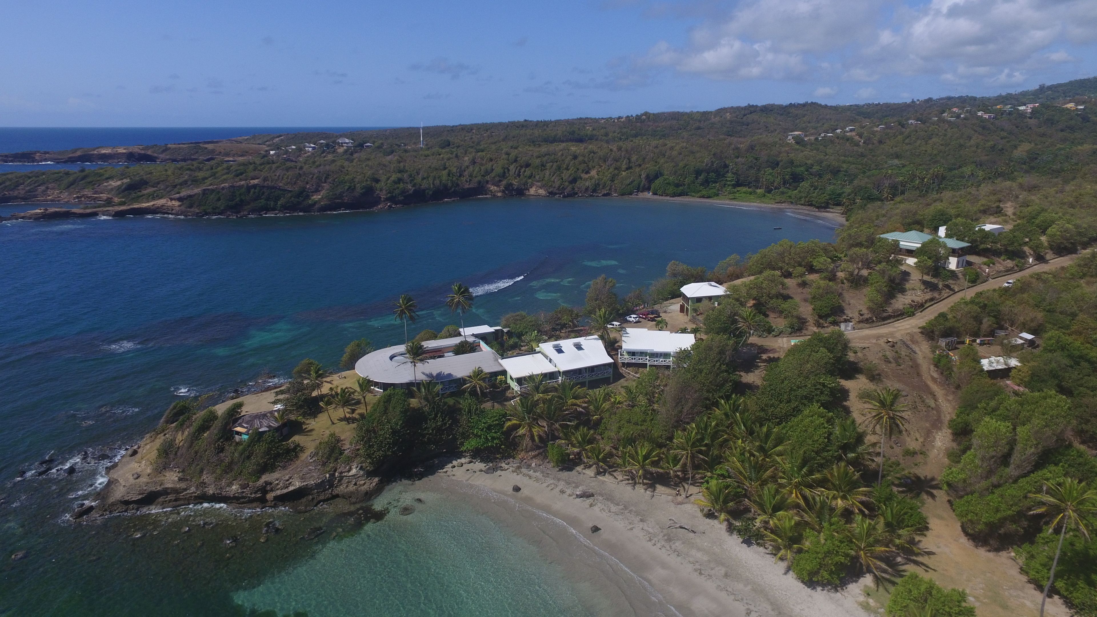 Vue aérienne d’une propriété située sur la côte, avec une plage, des eaux cristallines et une colline boisée.