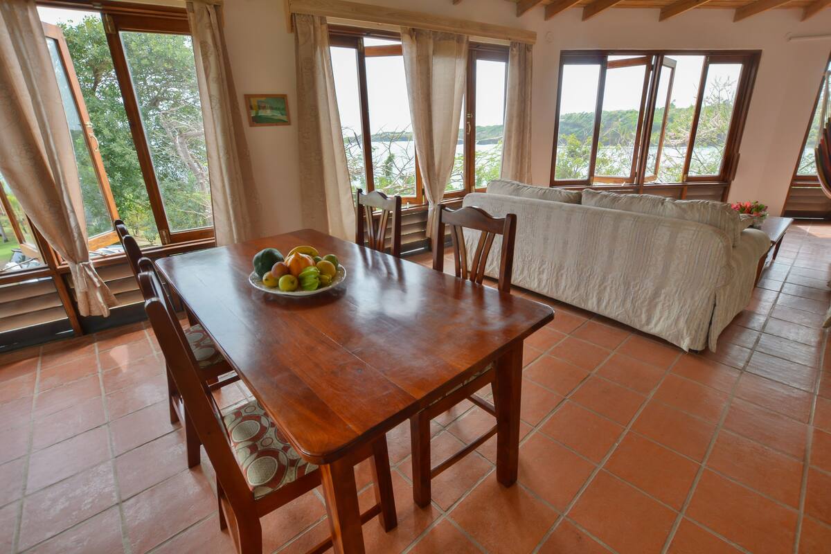 Uma área de refeições com uma mesa de madeira, cadeiras e uma tigela de frutas. Há um sofá e uma janela com vista para árvores e colinas.