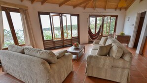 A spacious living area with two sofas, a coffee table, and a hammock. Large windows provide a view of the outdoors.