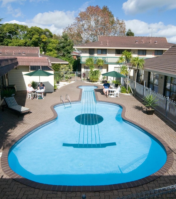 A swimming pool with a guitar-shaped design, surrounded by lounge chairs and umbrellas, with buildings in the background.