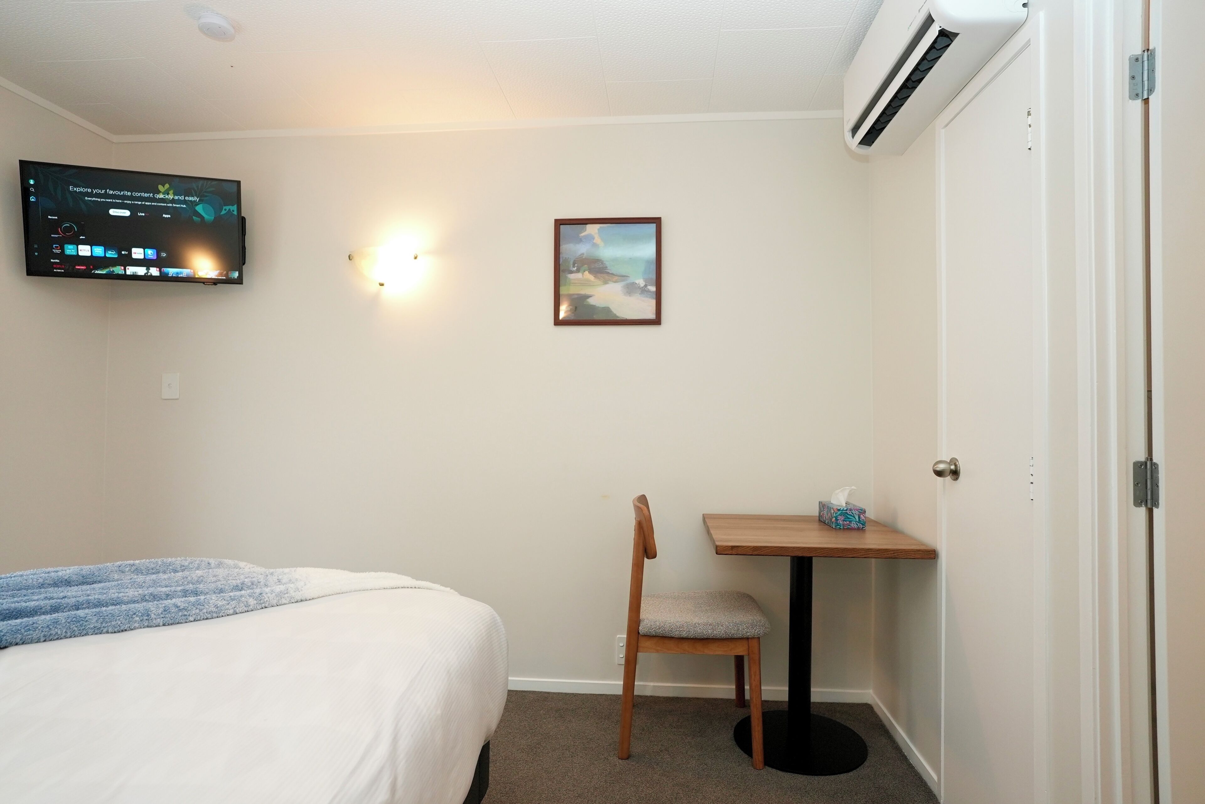 Habitación de hotel con cama, escritorio con silla, televisor montado y un cuadro en la pared.