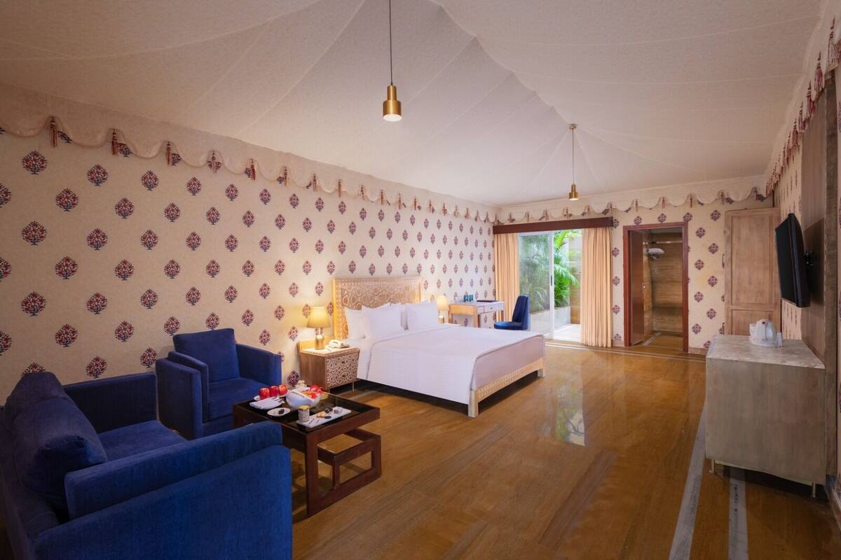 A hotel room with a bed, two blue armchairs, a wooden coffee table with breakfast items, a nightstand with a lamp, and a TV on the wall.