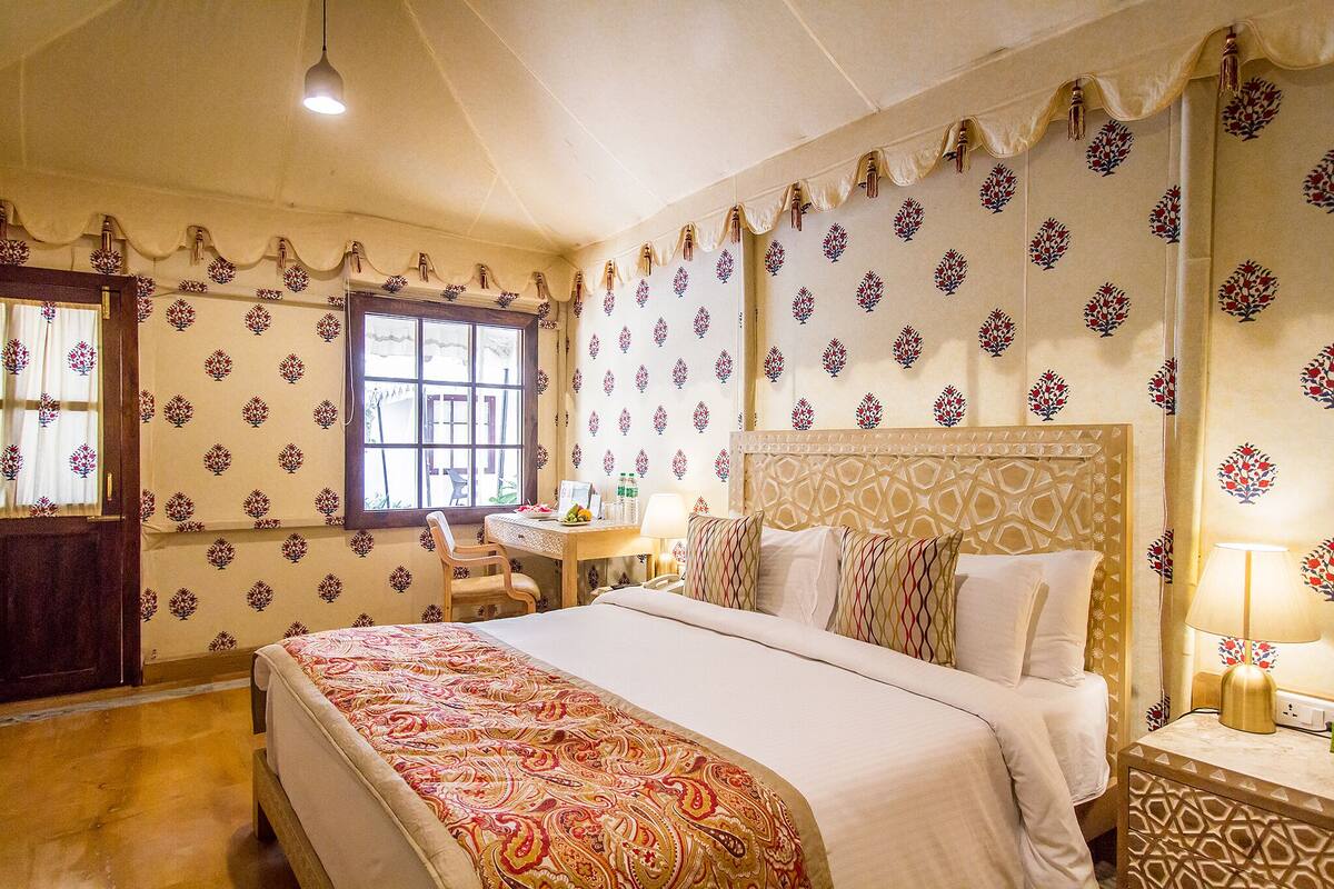 A hotel room with a large bed, a desk with a chair, a window, and patterned wallpaper.