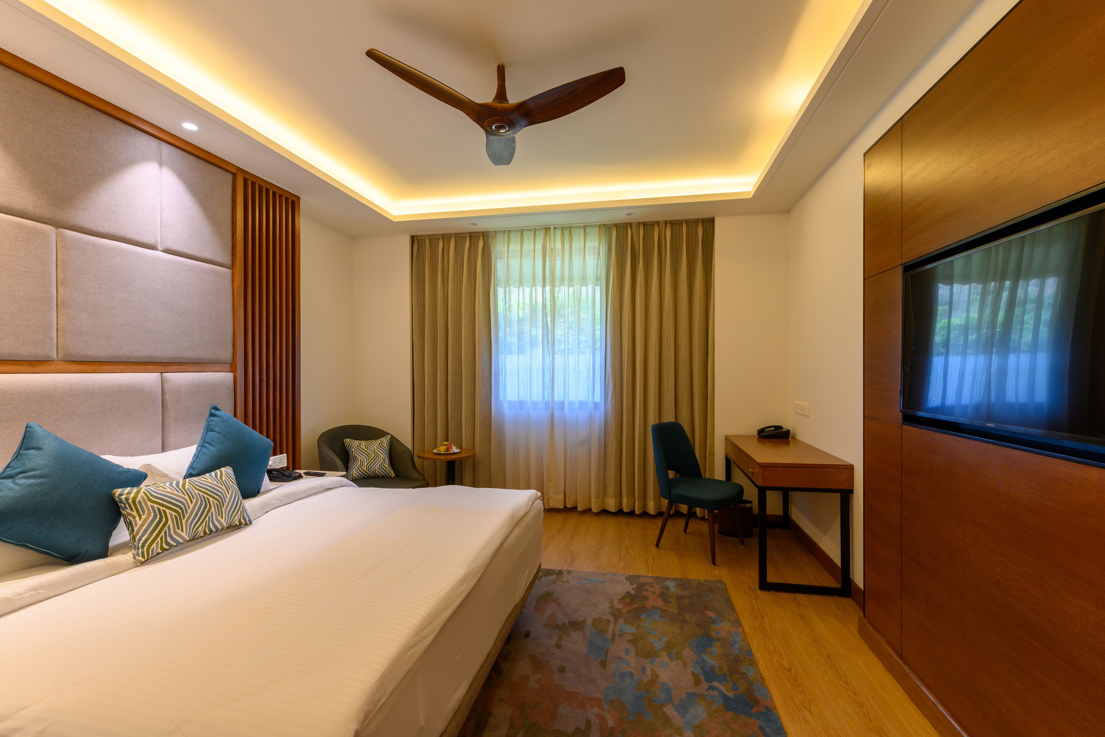 A hotel room with a large bed, a desk, a chair, a ceiling fan, and a TV.