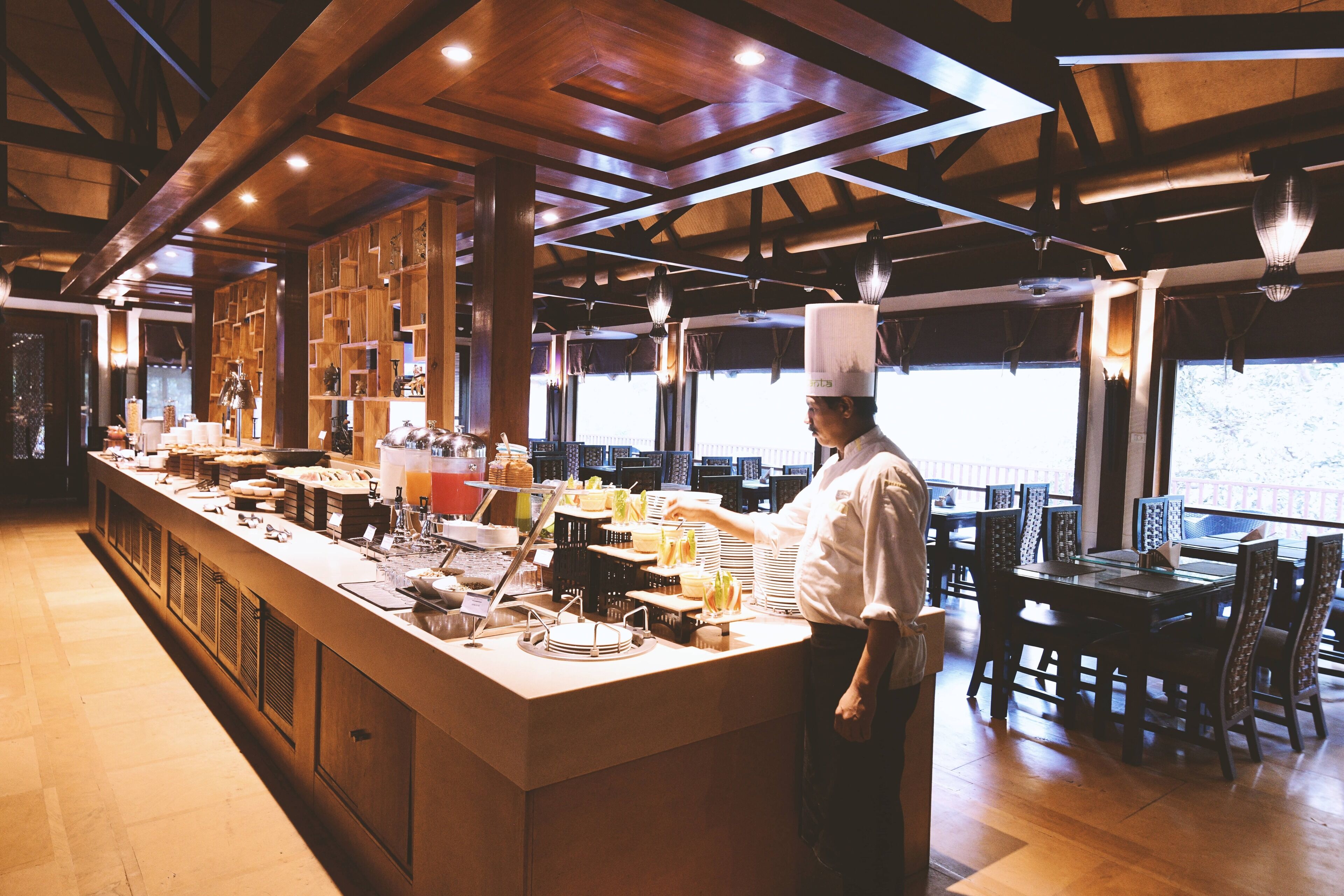 A chef in a kitchen with wooden beams and a long counter displaying various food items.