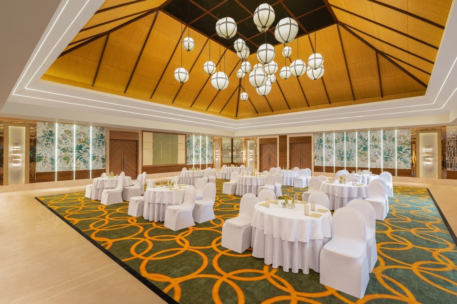 A spacious banquet hall with round tables and white chairs, a patterned carpet, and a high ceiling with hanging spherical lights.