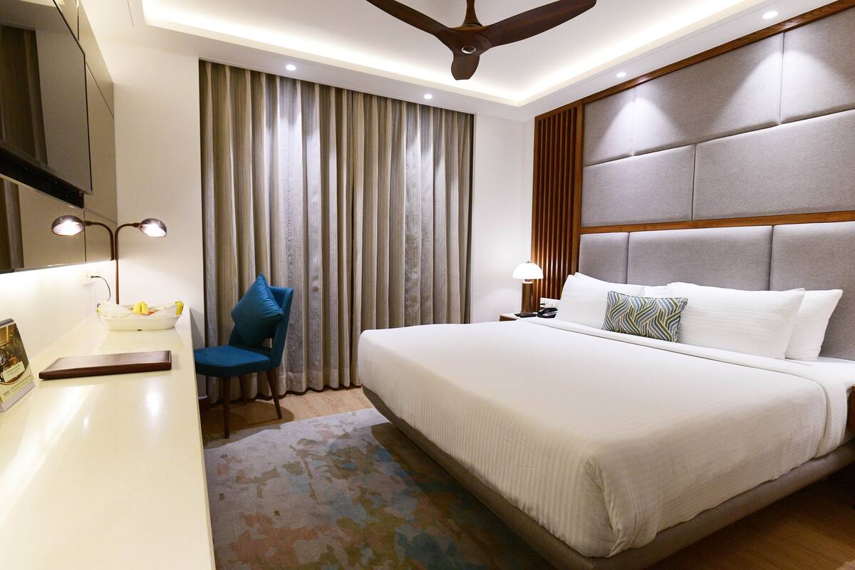 A hotel room with a large bed, a desk with a lamp, a chair, and a ceiling fan.
