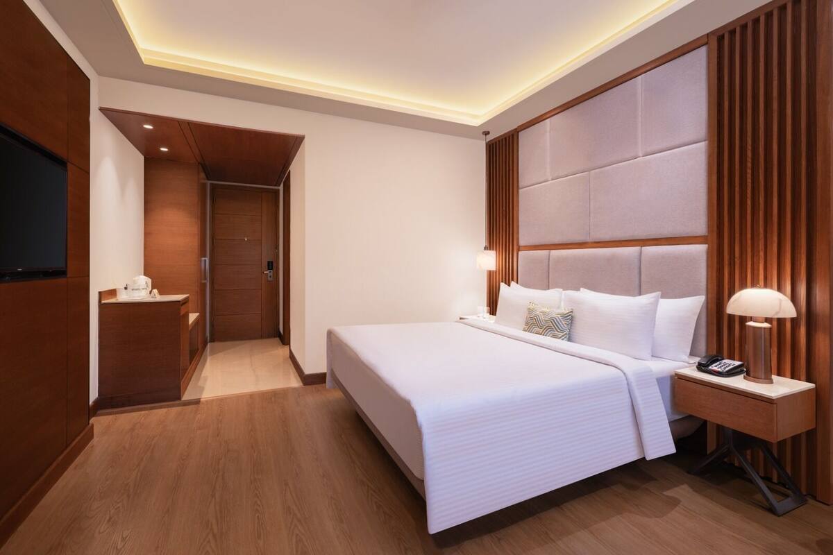 A modern hotel room with a large bed, a bedside table with a lamp, a TV mounted on the wall, and a wooden floor.