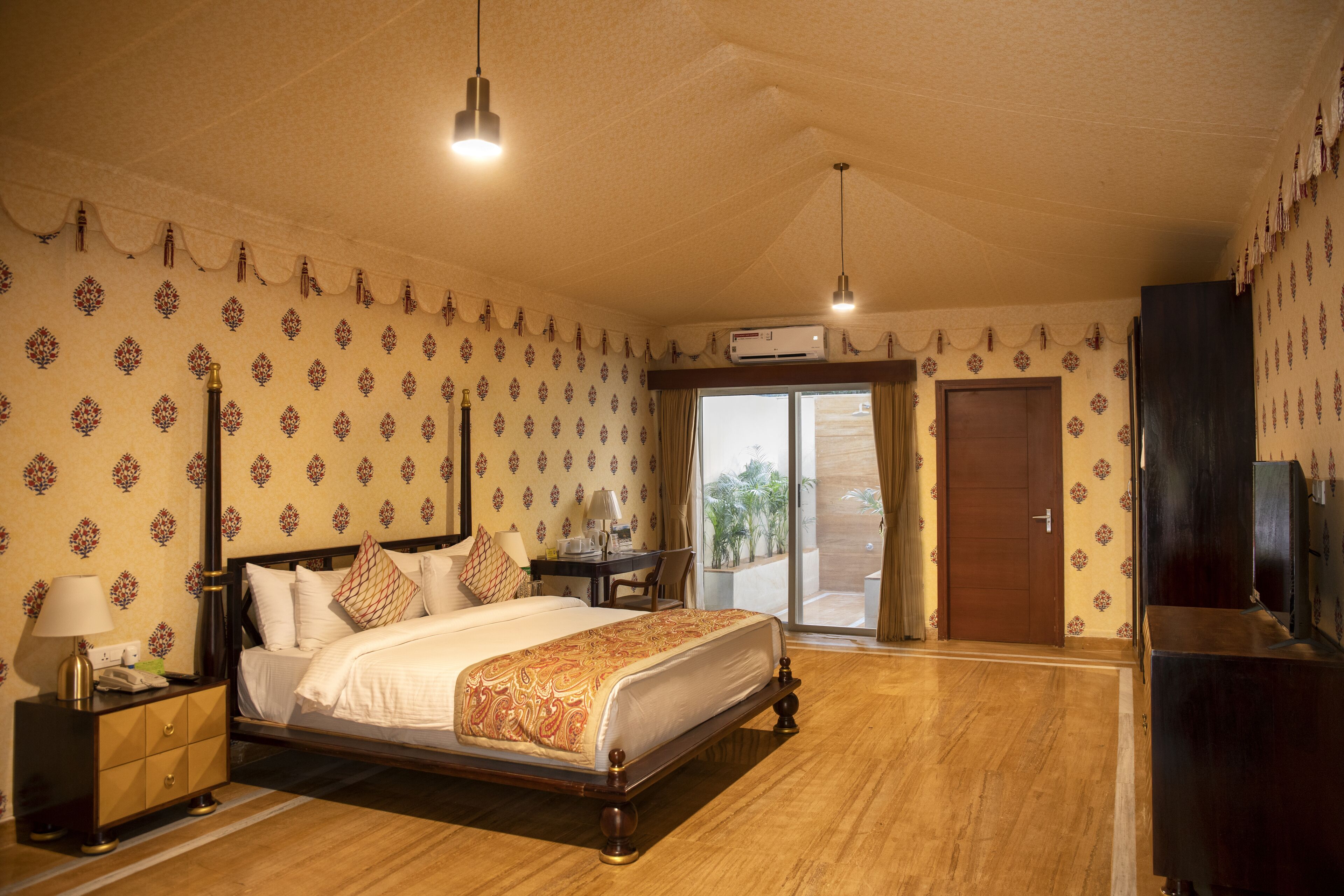 A hotel room with a bed, bedside table, lamp, and a desk with a chair. There is a door leading to a balcony.