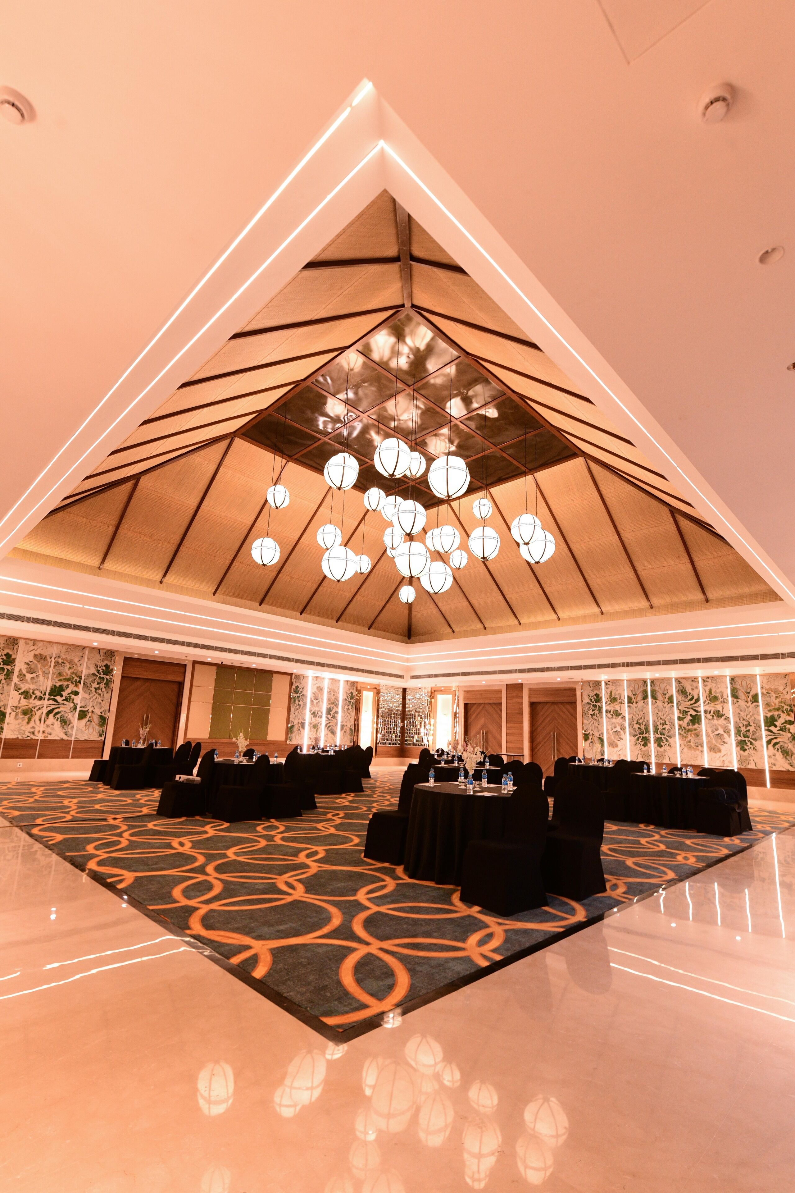 A spacious hall with a high ceiling, chandeliers, and a patterned carpet. Tables and chairs are set up for dining.