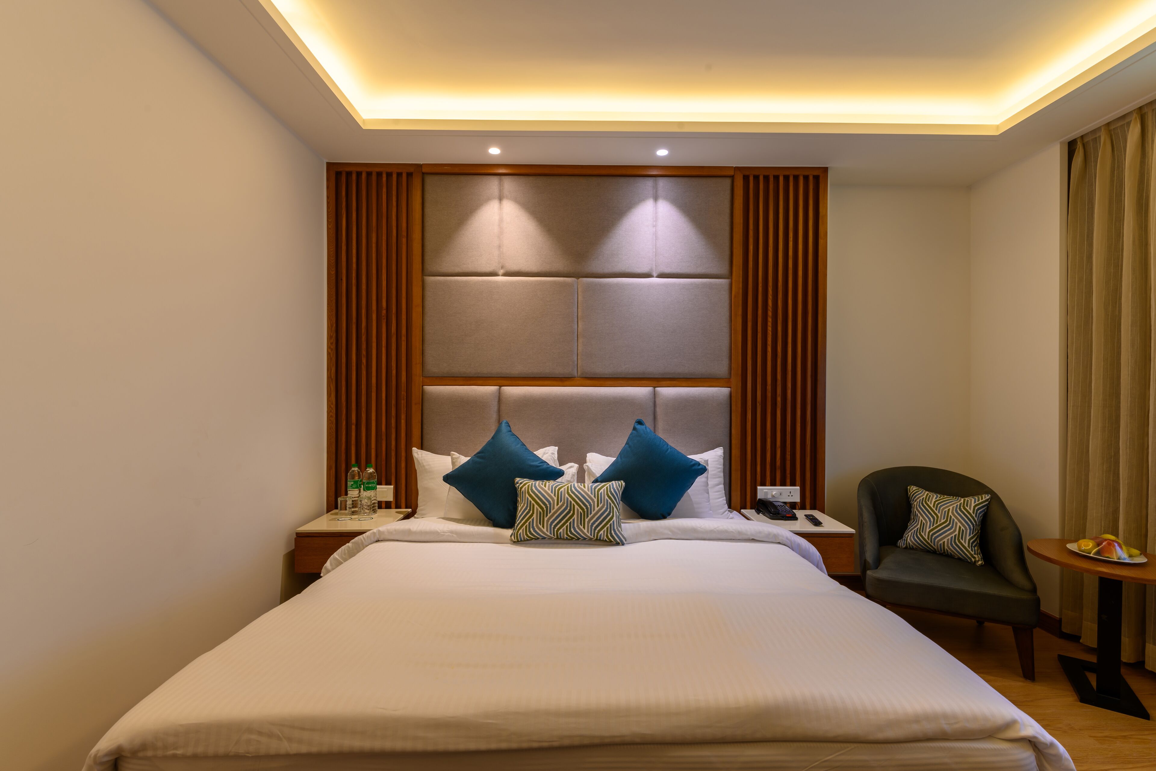 A hotel room with a large bed, a nightstand, a chair, and a small table with fruit.