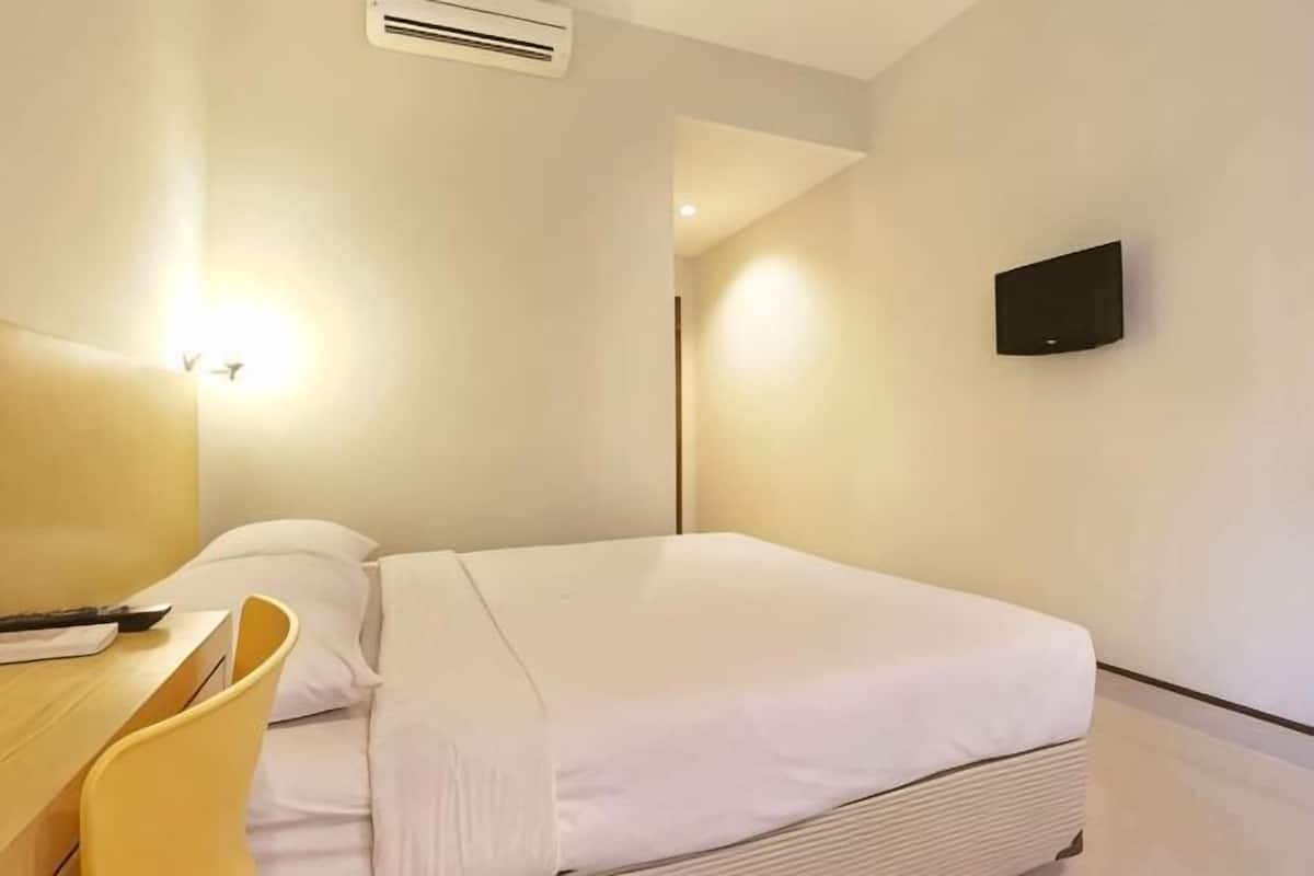 A hotel room with a bed, a desk with a chair, a flat-screen TV mounted on the wall, and an air conditioning unit.